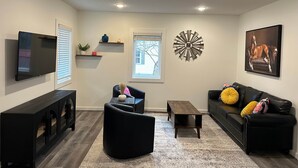 Una sala de estar con televisor de pantalla plana, sofás negros, una mesita de centro, un elemento decorativo en la pared y estanterías empotradas.