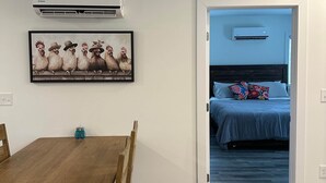 Una habitación con una mesa de comedor de madera, una obra de arte enmarcada de gallinas, un equipo de aire acondicionado y una puerta que conduce a un dormitorio con una cama y almohadas.