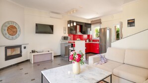 A modern living room with a sofa, a coffee table with a vase of flowers, a TV mounted on the wall, and a kitchen area with red cabinets and a stainless steel refrigerator. - Spa Villa Sunny in Chaniotis (Chaniotis)