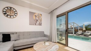 A modern living room with a grey sofa, a round marble coffee table, a wall clock, and a large glass door leading to a pool area. - Villa Lux Stana II in Zakynthos (Laganas)