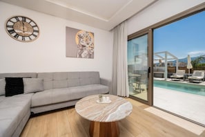 Ein modernes Wohnzimmer mit einem grauen Sofa, einem runden Marmor-Couchtisch, einer Wanduhr und einer großen Glasschiebetür zum Poolbereich.