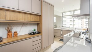 A modern kitchen with wooden cabinets, a marble backsplash, and a large window providing natural light. - Apartamento Primavera (Rio de Janeiro)