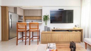 A modern kitchen with wooden cabinets, a built-in microwave, and a stainless steel refrigerator. There are two wooden chairs and a dining table. A flat-screen TV is mounted on the wall above a wooden console table. - Apartamento Primavera (Rio de Janeiro)