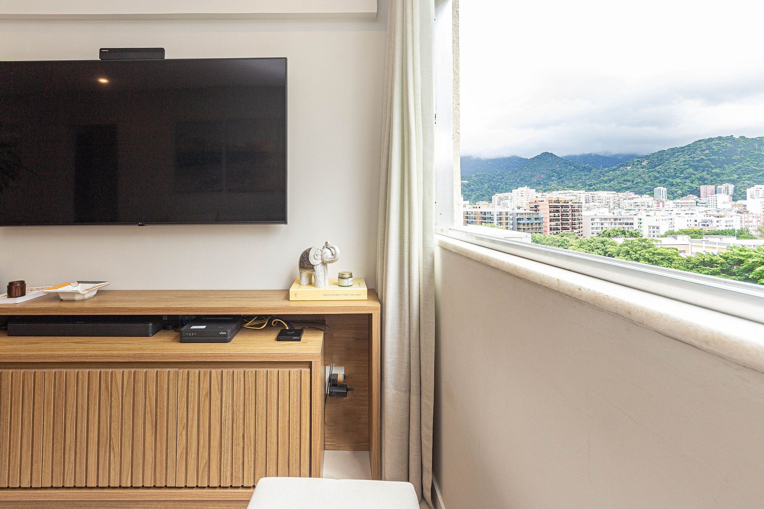 A room with a wooden desk, a flat-screen TV mounted on the wall, and a window offering a city view with mountains in the background.