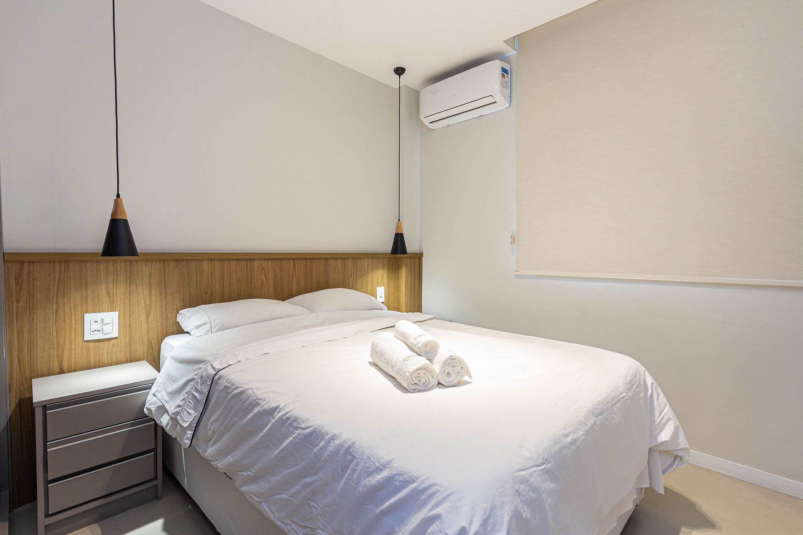 Una cama bien hecha con sábanas blancas y toallas enrolladas, una mesita de noche con cajones, una lámpara colgante y un equipo de aire acondicionado en la pared.