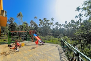 A playground with a slide and swings, surrounded by a green fence and palm trees.