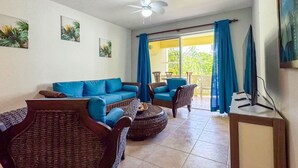Ett vardagsrum med en blå soffa, flätade stolar, en takfläkt och utsikt över grönskan genom fönstret.