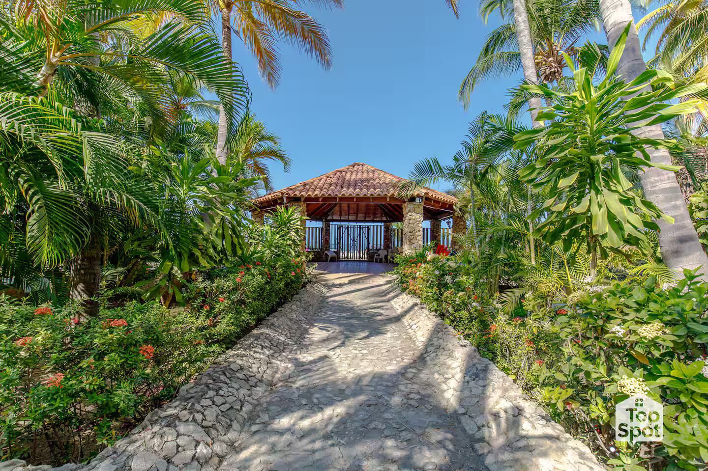 A stone pathway leading to a rustic building with a tiled roof, surrounded by lush tropical vegetation.