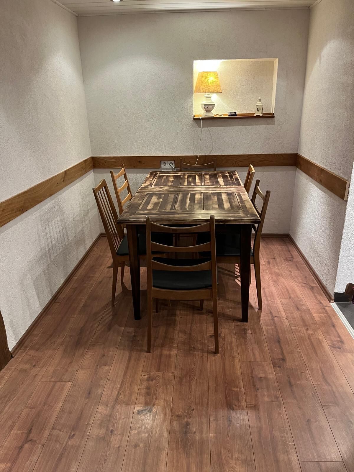 A dining area with a wooden table and chairs, a wall-mounted lamp, and a wooden floor.