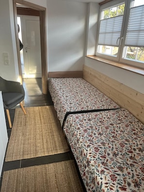 A small, clean room with two single beds, a chair, and a window with blinds.