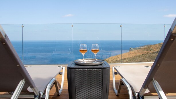 En balkon med to liggestole og et bord med to vinglas, med udsigt over havet.