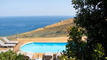 A pool with lounge chairs, a view of the ocean, and a hillside.