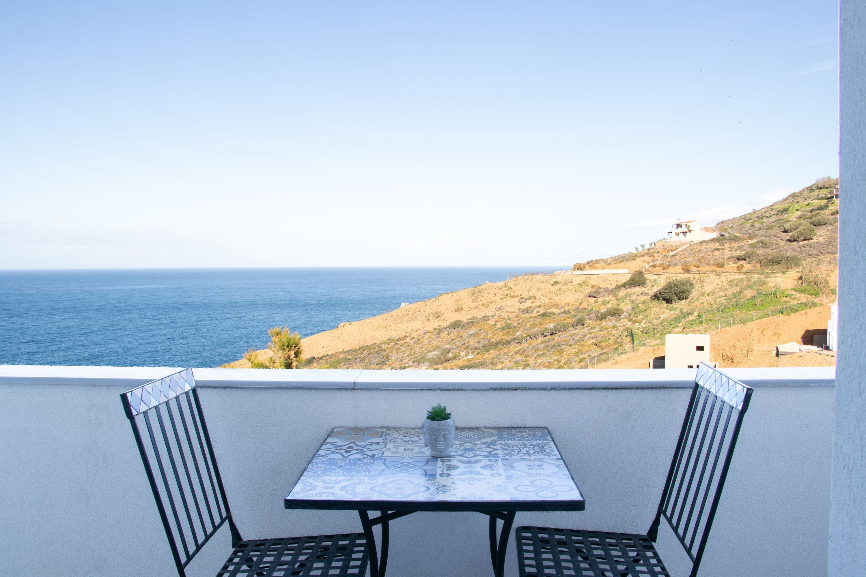 A balcony with a table and two chairs, overlooking a coastal landscape with a clear view of the sea and a hilly terrain in the background.