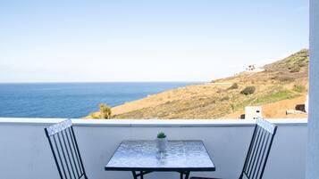 A balcony with a table and two chairs, overlooking a coastal landscape with a clear view of the sea and a hilly terrain in the background.