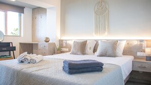 A modern hotel room with a large bed, a bedside table, a nightstand, a wall-mounted macrame decor, and a window with a view. - House of Harmony Suites | Maria (Agia Pelagia)