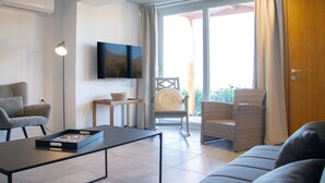 A modern living room with a sofa, a coffee table, a TV mounted on the wall, and a sliding glass door leading to an outdoor area. - House of Harmony Suites | Maria (Agia Pelagia)