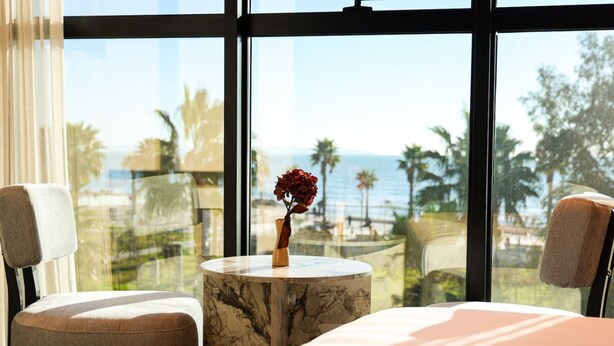 Ein Schlafzimmer mit einem Bett, zwei Stühlen, einem мраморный Beistelltisch und einer Vase mit roten Blumen. Ein großes Fenster bietet Blick auf Palmen und das Meer.