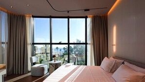 Ein modernes Hotelzimmer mit einem großen Bett, einer Sitzecke mit Tisch und Stühlen sowie Blick ins Freie durch große Fenster.