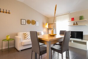 A modern living room with a dining table, two chairs, a sofa, a small side table, a lamp, a TV, and wall decorations. - Desenzano Suites - Tiglio by Wonderful Italy (Desenzano del Garda)