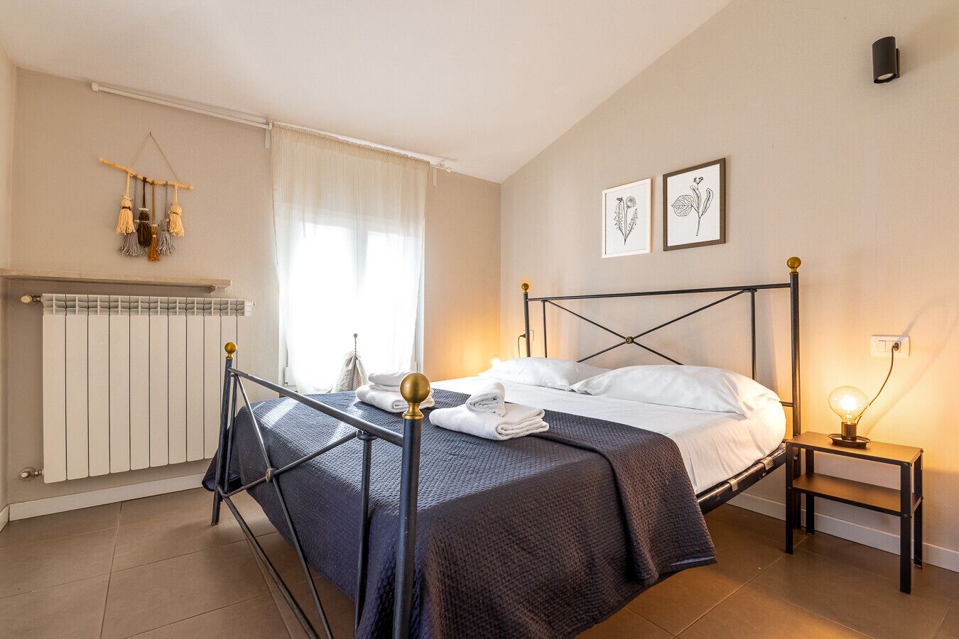 Ein Schlafzimmer mit Bett, Heizkörper, Nachttisch mit Lampe und Fenster mit Vorhängen.