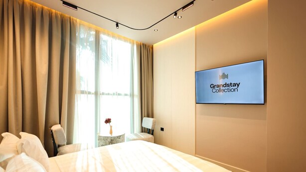 Ein Hotelzimmer mit einem Bett, einem Fernseher, der 'Grandstay Collection' zeigt, einem Fenster mit Vorhängen und einem kleinen Tisch mit einer Vase.