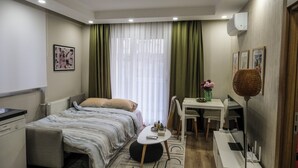 A modern hotel room with a bed, a desk, a chair, a small table, a radiator, and a window with curtains. - Smartly Designed Cozy Central 1BR Apartment (Antalya)