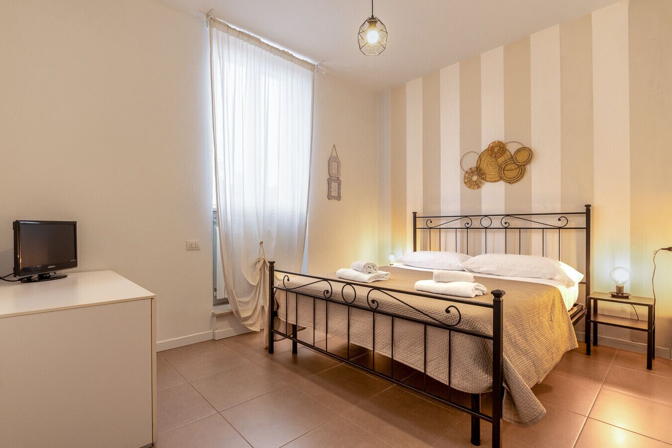 Un dormitorio con cama, mesita de noche, televisor y ventana con cortinas.
