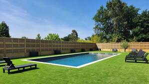 A rectangular swimming pool with a surrounding grassy area, two lounge chairs, and a wooden fence.