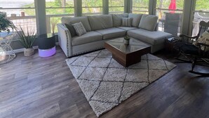 Amplia sala de estar con sofá seccional, una mesa de centro y ventilador de techo. Ventanas grandes ofrecen vistas al exterior.