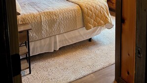 Un dormitorio de cabaña de madera con una cama, una ventana con cortinas, una lámpara fijada a la pared y un cuadro enmarcado.