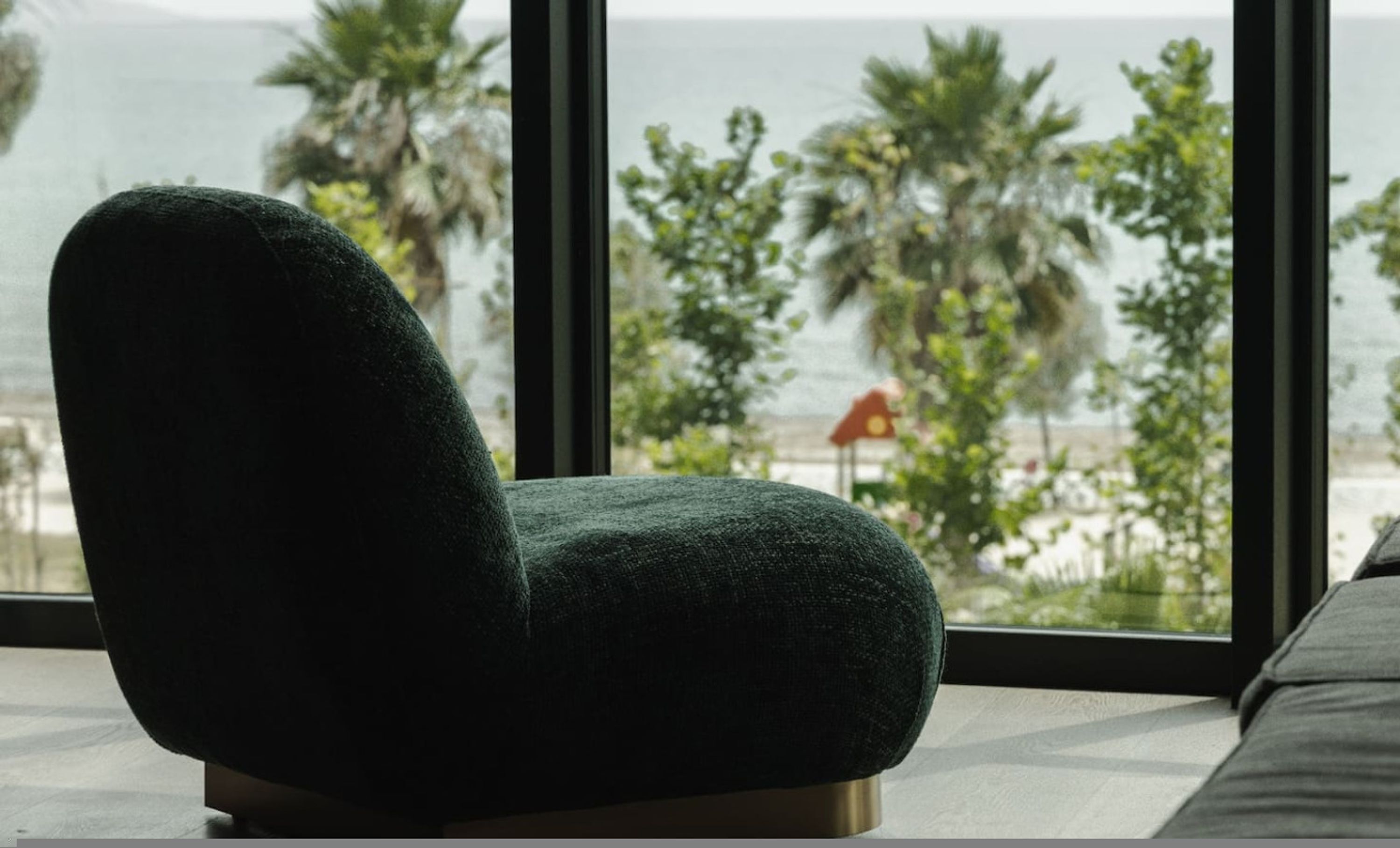 A dark green upholstered chair with a metallic base, positioned in front of a large window with a view of palm trees and a beach.