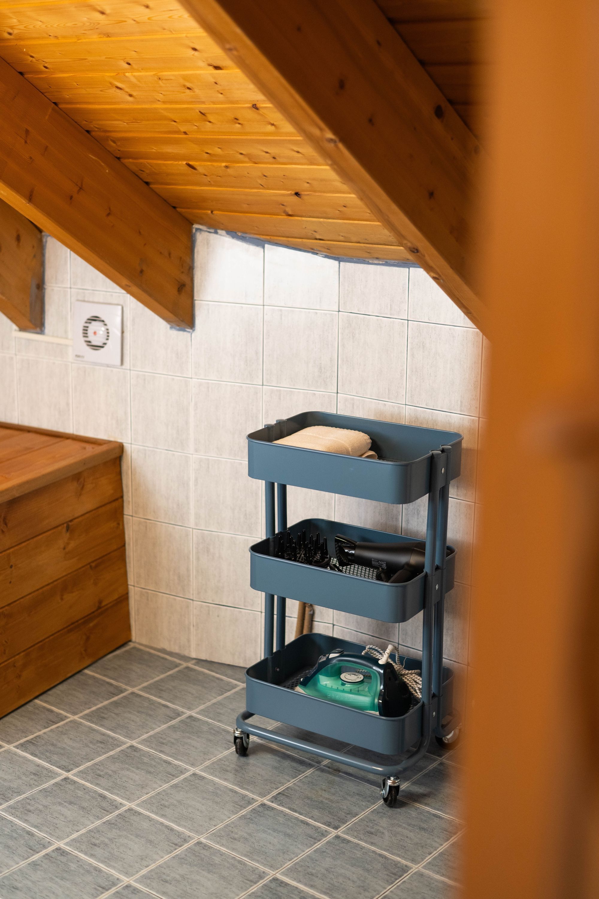 A corner of a room with a wooden ceiling, a tiled wall, and a blue storage cart with items on it.