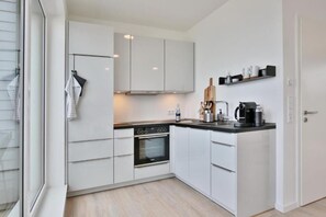 Une cuisine moderne avec des armoires blanches, un four encastré, une machine à café et une étagère en bois où sont rangés des ustensiles de cuisine.
