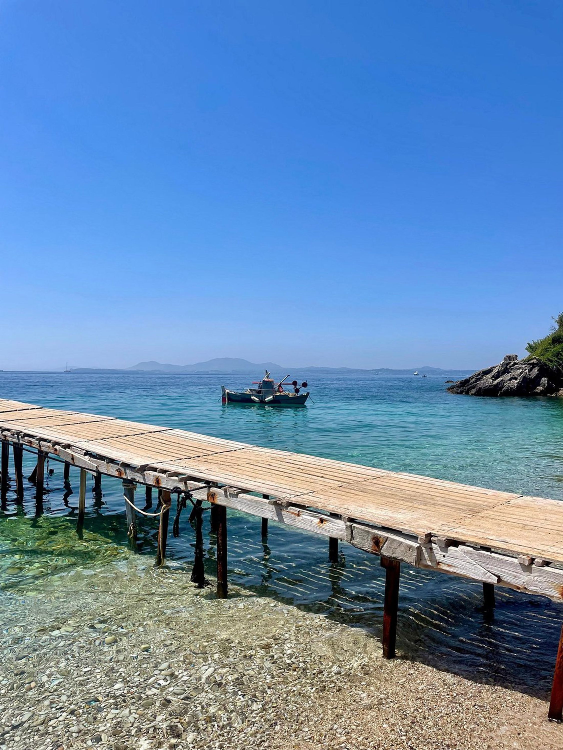 A wooden pier extending into clear blue water with a boat anchored in the distance.