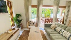 Una sala de estar con sofá, una mesa de centro, televisión y un comedor visible a través de puertas de vidrio.