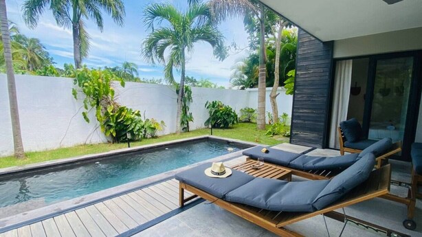 A modern outdoor pool area with wooden deck flooring, a lounge chair with cushions, and a glass door leading to a covered patio. - Private Pool Villa | Parking | Washer (Las Terrenas)