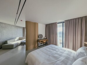 Habitació d'hotel moderna amb un llit gran, una taula de treball amb una cadira, un sofà i vista a l'exterior a través de grans finestres.