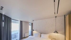 Ein modernes Hotelzimmer mit einem großen Bett, einem Nachttisch mit Lampe, Blick auf die Stadt durch große Fenster und einem Balkon.