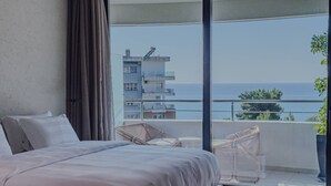 Een slaapkamer met een groot bed, een balkon met rieten meubilair en uitzicht op zee en gebouwen.