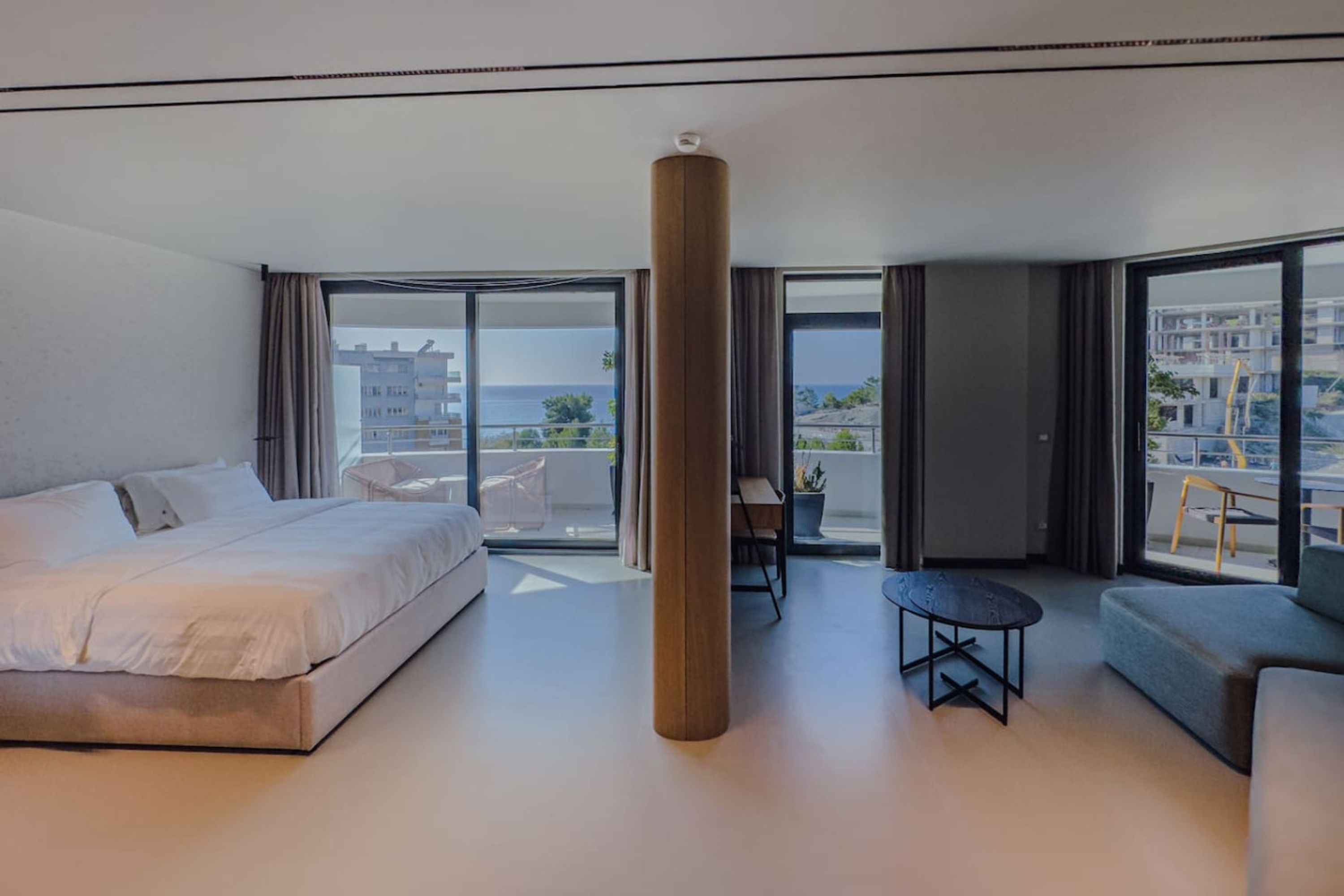 A modern hotel room with a large bed, a sofa, a small table, and a view of the sea through large windows.