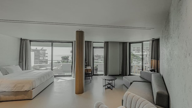 Een moderne hotelkamer met een groot bed, een zithoek met een bank en fauteuils, een kleine tafel en uitzicht op de stad door grote ramen.
