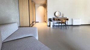 Ein modernes, minimalistisches Zimmer mit grauem Sofa, einem Holzschrank, einem Schreibtisch mit Stuhl, einem runden Spiegel und einem weißen Heizkörper.