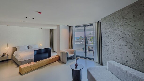 Ein modernes Hotelzimmer mit einem Bett, einer Couch, einem Fernseher auf einem Holzboden, einem kleinen Tisch mit einer Vase und einem Balkon mit Blick.