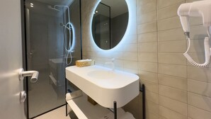 Ein modernes Badezimmer mit Duschkabine aus Glas, einem runden Spiegel, einem weißen Waschbecken und einem an der Wand befestigten Haartrockner.