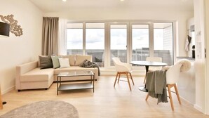 Ein modernes Wohnzimmer mit einer Couch, einem Couchtisch und Sesseln, großen Fenstern und einem Balkon.