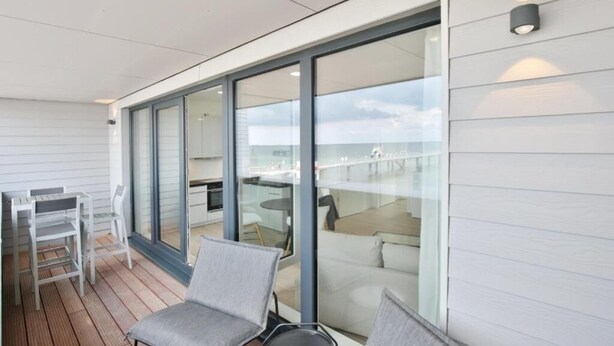 Eine moderne Terrassenanlage mit Glaswand, Essbereich und Sitzgruppe mit Tisch und Stühlen.