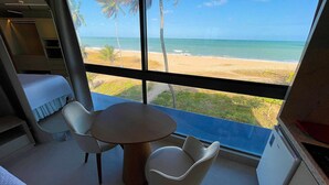 Ein Zimmer mit Blick auf Strand und Meer durch große Fenster. Es gibt einen runden Tisch mit zwei Stühlen und eine kleine Küchenzeile.