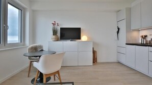 Moderni keittiö, jossa on valkoiset kaapit, tasokiinnitetty litteänäyttöinen televisio, ruokailutila lasipöydällä ja valkoisilla tuoleilla sekä ikkuna, josta tulee luonnonvaloa.