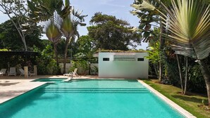 A rectangular swimming pool with clear blue water, surrounded by lush greenery and a small white building in the background. - 2 Bedroom Pool Apartment (Las Terrenas)