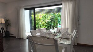 A dining area with a glass table set for four, white chairs, and a view of greenery through a large window. - 2 Bedroom Pool Apartment (Las Terrenas)